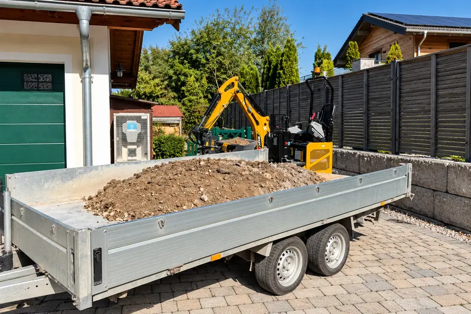 Minibagger hinter einem Anhänger voller Aushub in einer gepflasterten Einfahrt