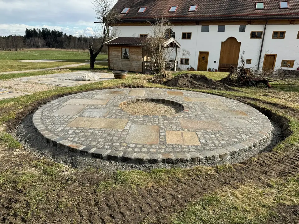 Runder Feuerstellen-Platz aus Granitpflaster mit Natursteinplatten und Kiesrund in der Mitte vor einem Bauernhaus